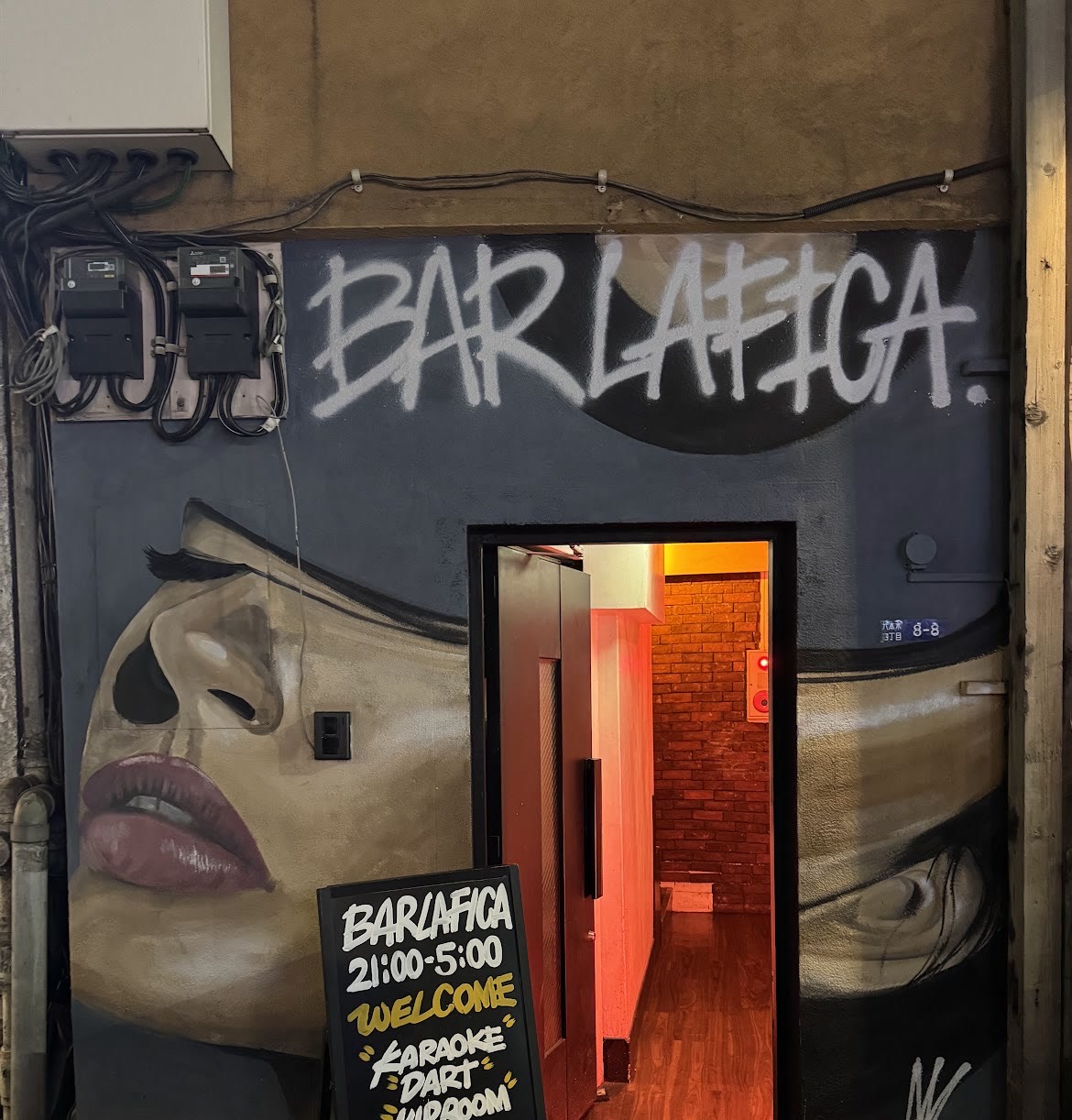 夜の「BAR LA FICA」の入口。灰青色の壁に女性の顔の壁画と白いスプレー文字「BAR LAFICA.」。開いた黒い扉の奥に赤い照明の通路とレンガ調の壁、手前に「BARLAFICA 21:00-5:00 WELCOME KARAOKE DART VIPROOM」と書かれた黒板の立て看板。