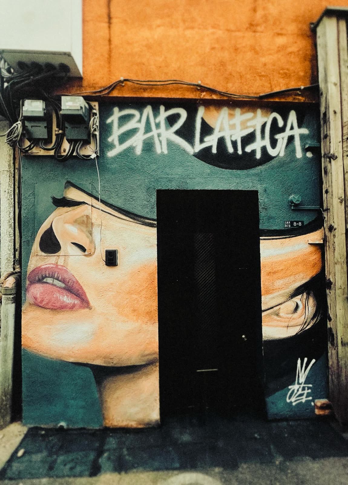 昼の「BAR LA FICA」の入口。女性の顔の壁画の中に黒い扉、上に白いスプレー文字「BAR LAFICA.」、扉の右に住居表示「六本木 四丁目 8-8」。